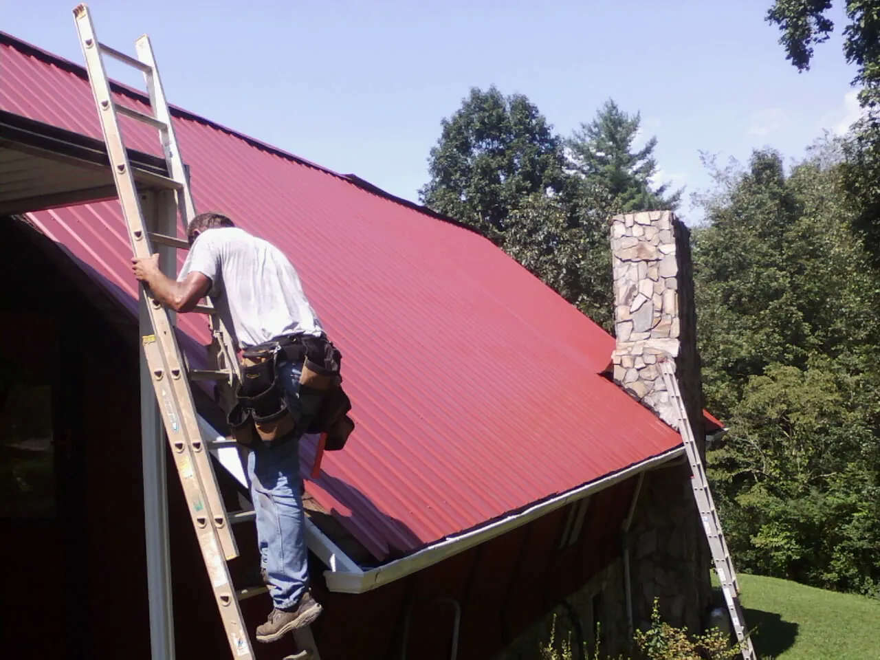 Roofer on a ladder beside a red metal roof and stone chimney — Soffit & Fascia Repair by Roofing Brothers in Cowlesville